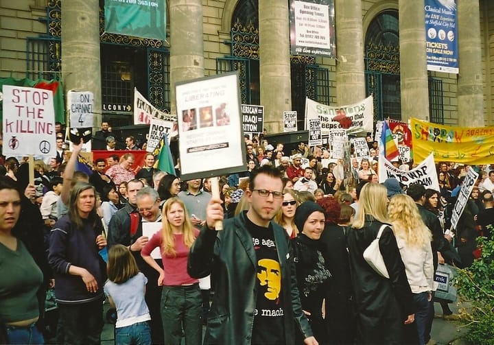 A person with dark hair and glasses in all black, with a RATM t-shirt and a placard, amongst dozens of other demonstrators
