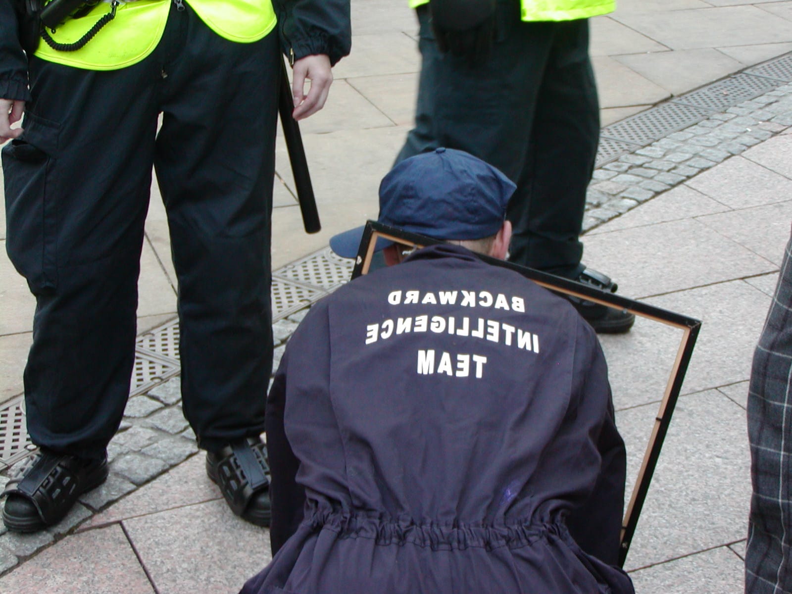 A protesting performance artist crawling in front of cops, wearing overalls that say "backward intelligence team" written in reverse, and a frame around their neck