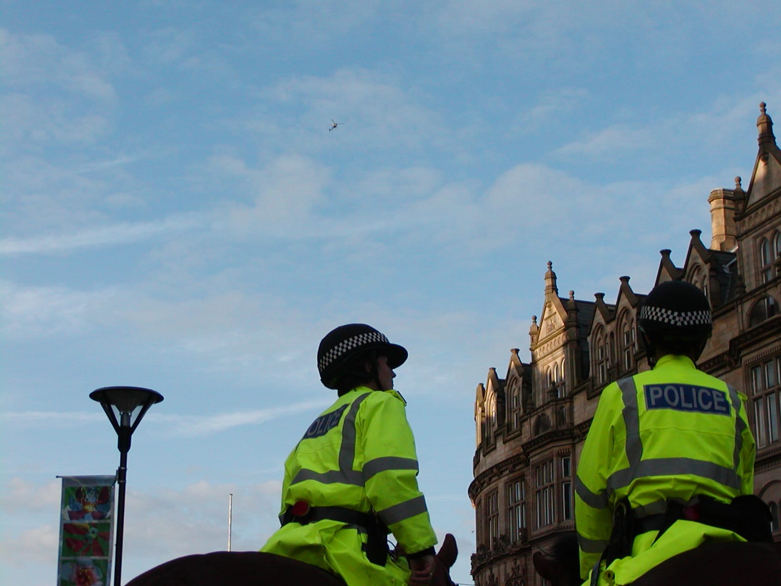 Two mounted police officers with old buildings in the background under a blue sky where a helicopter hovers