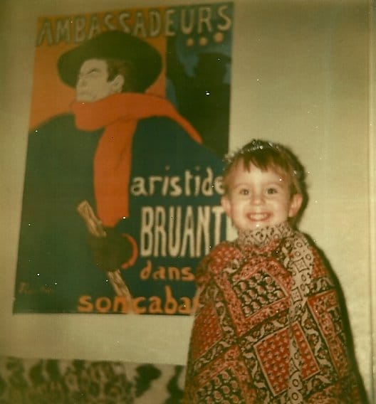 A toddler with blonde hair and blue eyes, wrapped in a throw, wearing tinsel, in front of a French poster.