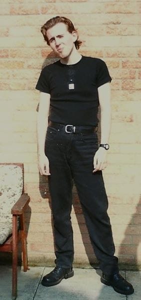A teenager in all black, with dark hair, standing awkwardly in front of a brick wall.