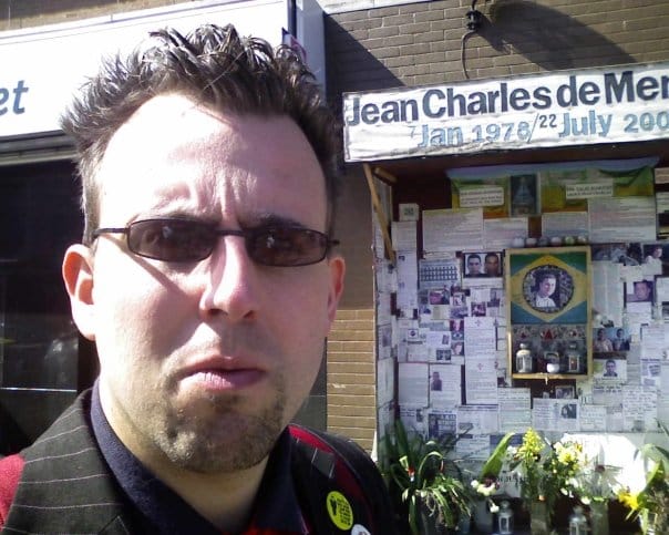 A person in a pinstripe jacket with pin badges and dark glasses, in front of the Jean Charles de Menezes memorial