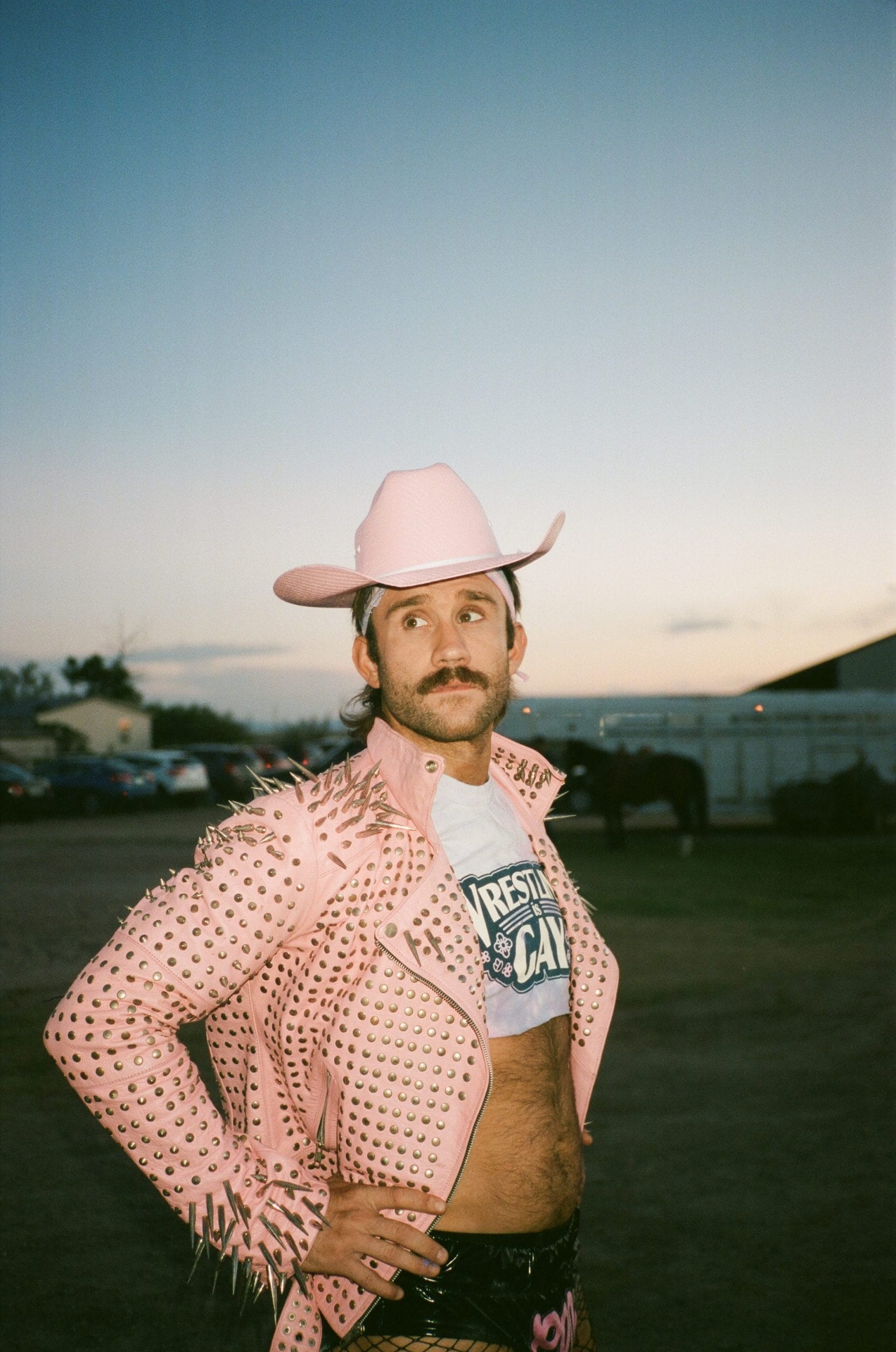 A mustachioed man in a cowboy hat, and a studded jacket over a crop top that states "wrestling is gay"