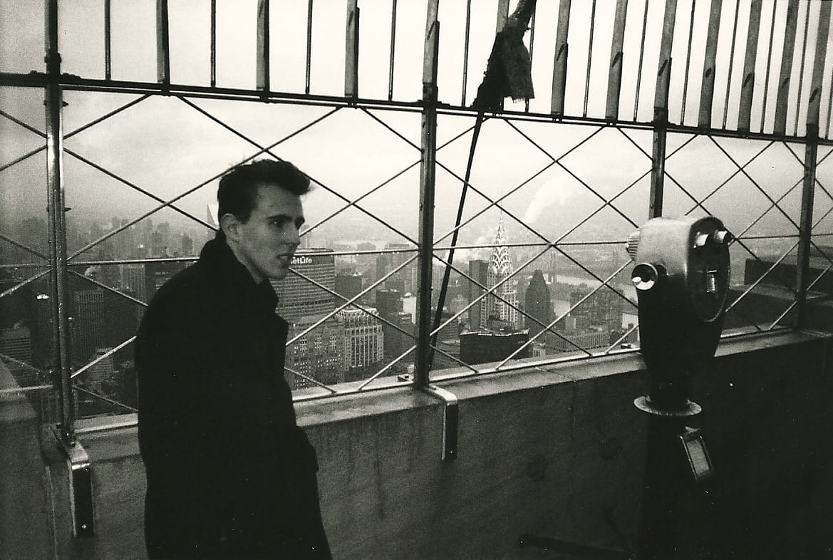 A person in a long black overcoat, standing aloof at the top of the Empire State Building, in black and white.